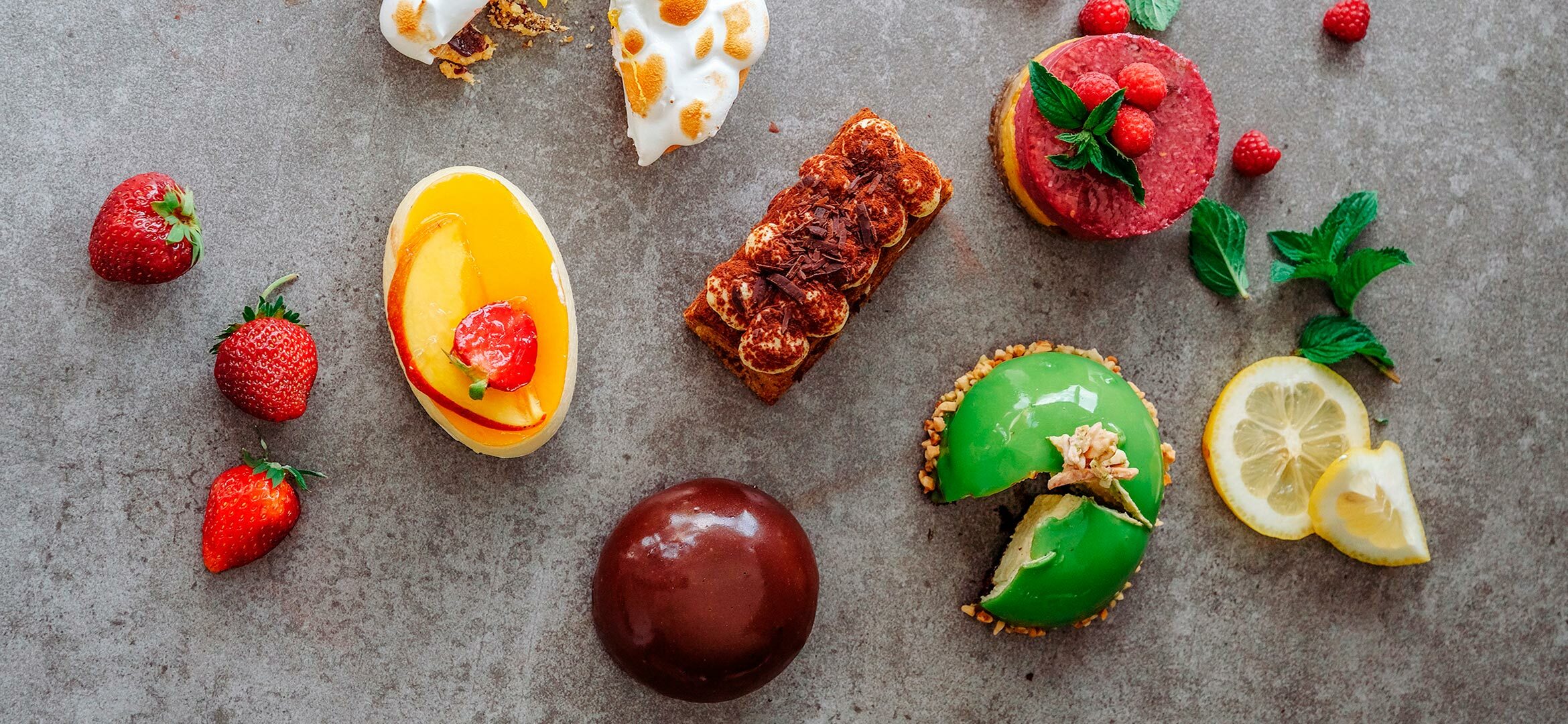 Blick auf Süßspeisen – Kuchen, Tiramisu, Schaumgebäck – die auf einer grauen Schieferplatte angerichtet sind. Dazwischen Erdbeeren und Minzblätter.