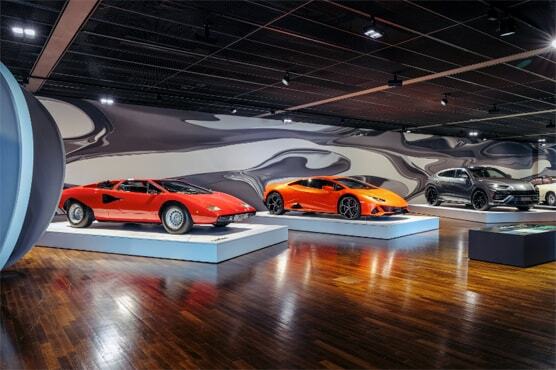 Foto in den Ausstellungsraum, in dem drei historische Lamborghinis nebeneinander präsentiert werden. An den Wänden sind organische grau-weiße Muster.
