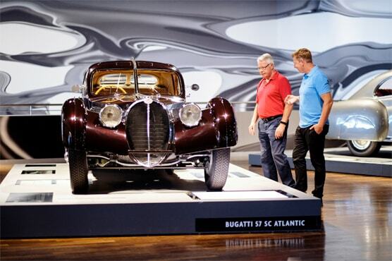 Foto von zwei weißen Menschen, die in einem Ausstelungsraum neben einem historischen Bugatti stehen und ihn betrachten, gestikulieren.