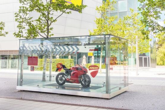 Foto eines roten Ducati Motorrades, dass in der Parklandschaft der Autostadt zwischen Bäumen in einem gläsernen Kasten-Display steht.
