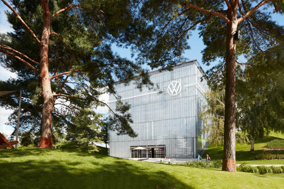 Blick aus der Parkanlage der Autostadt auf die Fasade des VW Pavillon. Zwischen Bäumen ragt das helle Gebäude mit großen Logo empor.