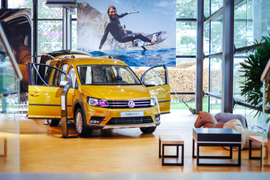 Foto eines gelben VW Nutzfahrzeug im Pavillon. Die Vordertüren sind geöffnet, es parkt vor einem Foto eines Surfers, neben einer Kinderecke.