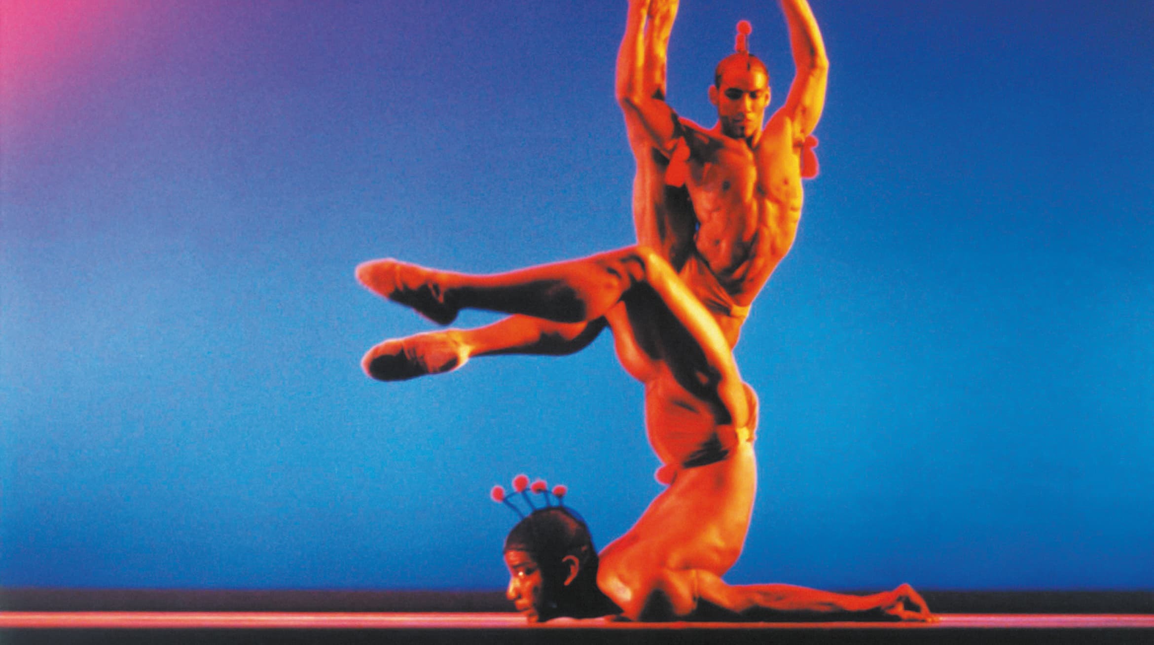 Foto von zwei Akrobaten auf einer Bühne vor blauem Hintergund.Die Personen sind sehr muskulös und befinden sich in komplexen Posen.