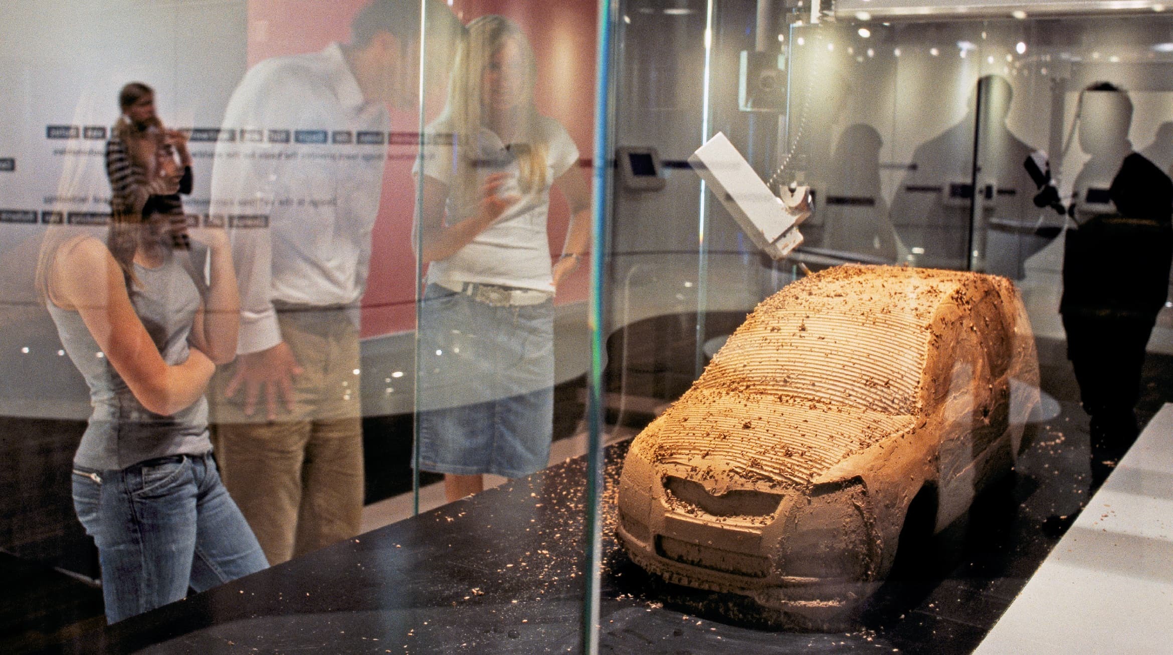 Foto eines Glaskasten-Displays. Darin befindet sich ein Automodell aus einem Ton-ähnlichen Material. Menschen stehen um diesen Kasten.
