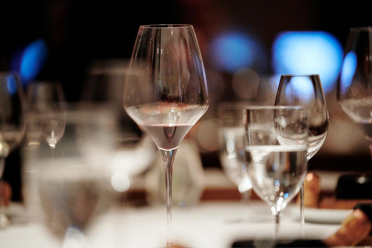 Foto von Weingläsern, die auf einem Tisch stehen. Manche sind unscharf, andere scharf. Mittig steht ein Weinglas, in dem sich etwas Rotwein befindet.