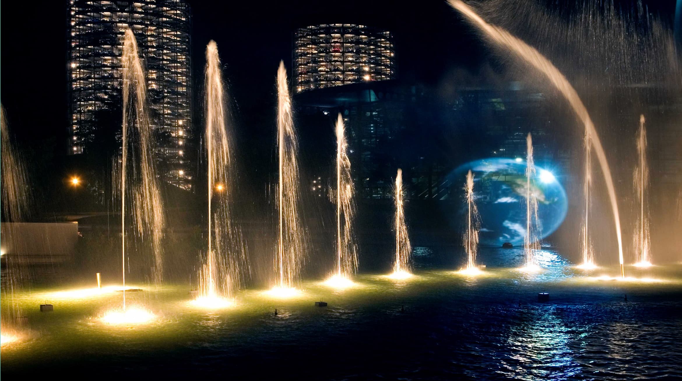 Foto einer Wassershow in der Autostadt. Beleutete Fontänen spritzen in der Nacht aus einem Gewässer. Im Hintergrund leuchten die beiden Abholtürme.