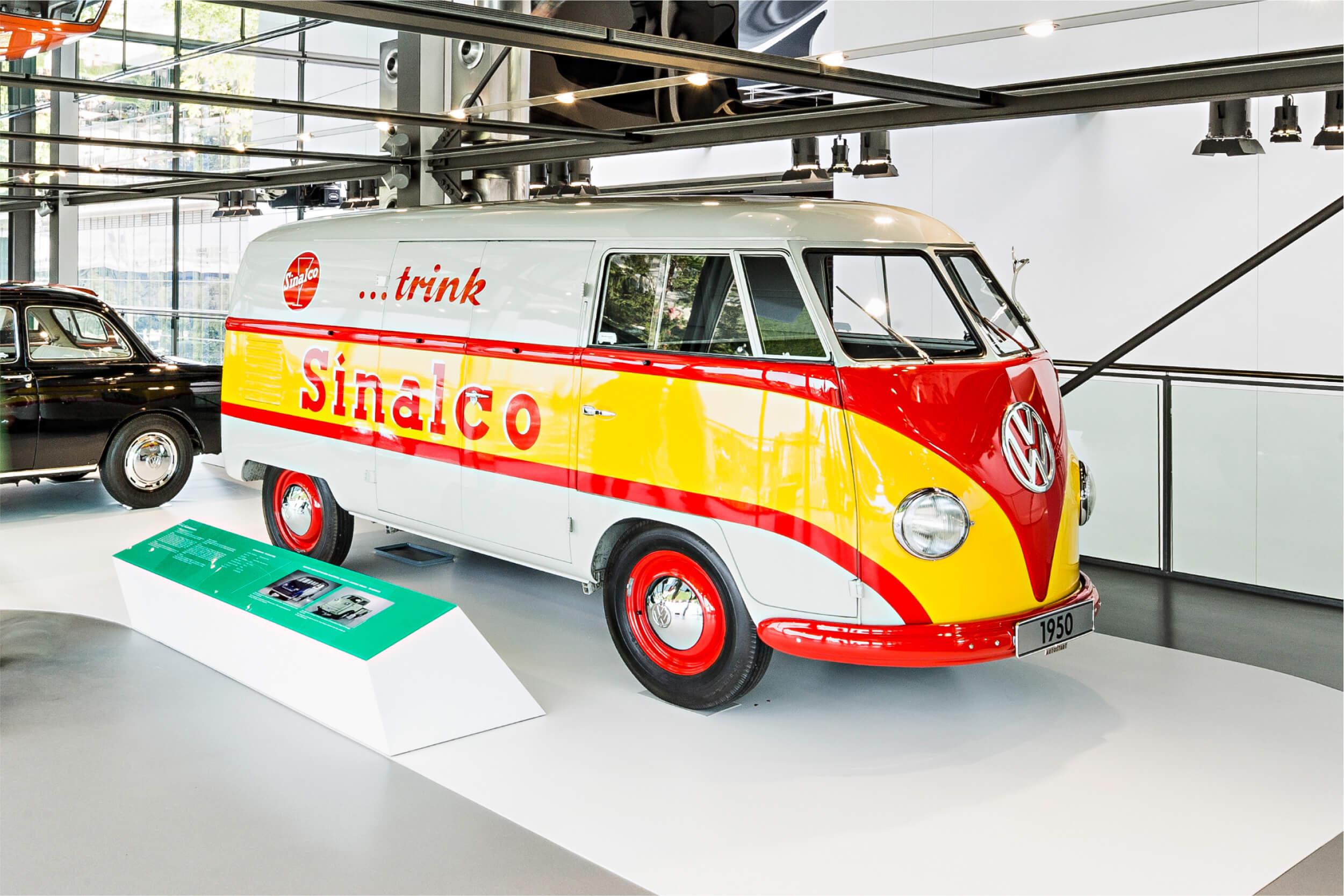 Seitenansicht eines historischen VW-Bulli Modells, der in einem Ausstellungsraum steht. Auf der Seite des Autos ist der Werbeslogen "..trink Sinalco".