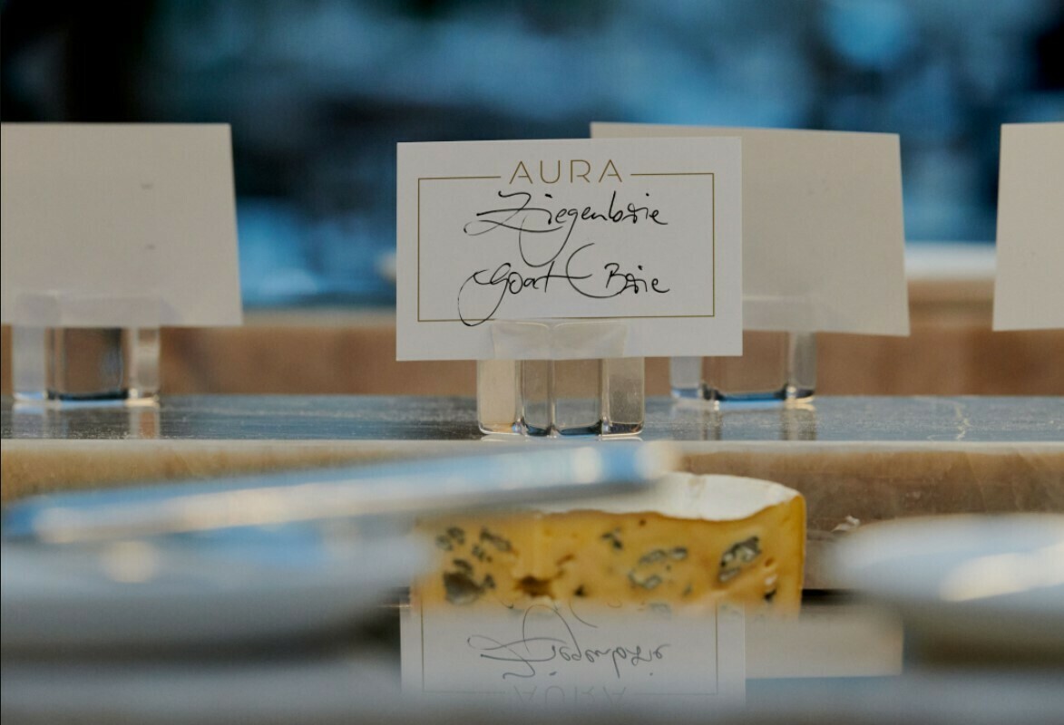 Nahaufnahme eines Käse. Hinter dem Stück Käse befindet sich ein Schild in Glashalterung. Darauf steht "AURA Ziegenbrie, goat brie".