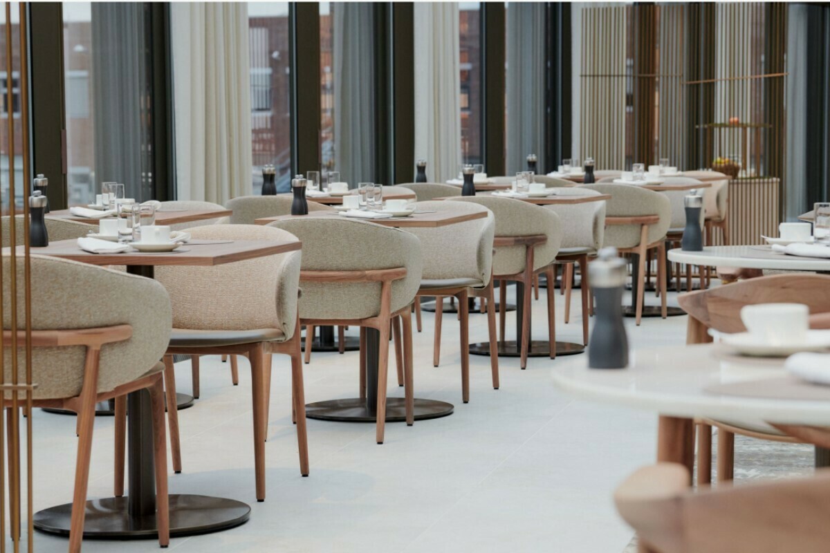 Foto der Frühstückslounge. Eine Reihe an Zweier-Tisch-Konstellationen mit Gedeck und beige-bepolsterten Holzstühlen an einer Fensterfront.