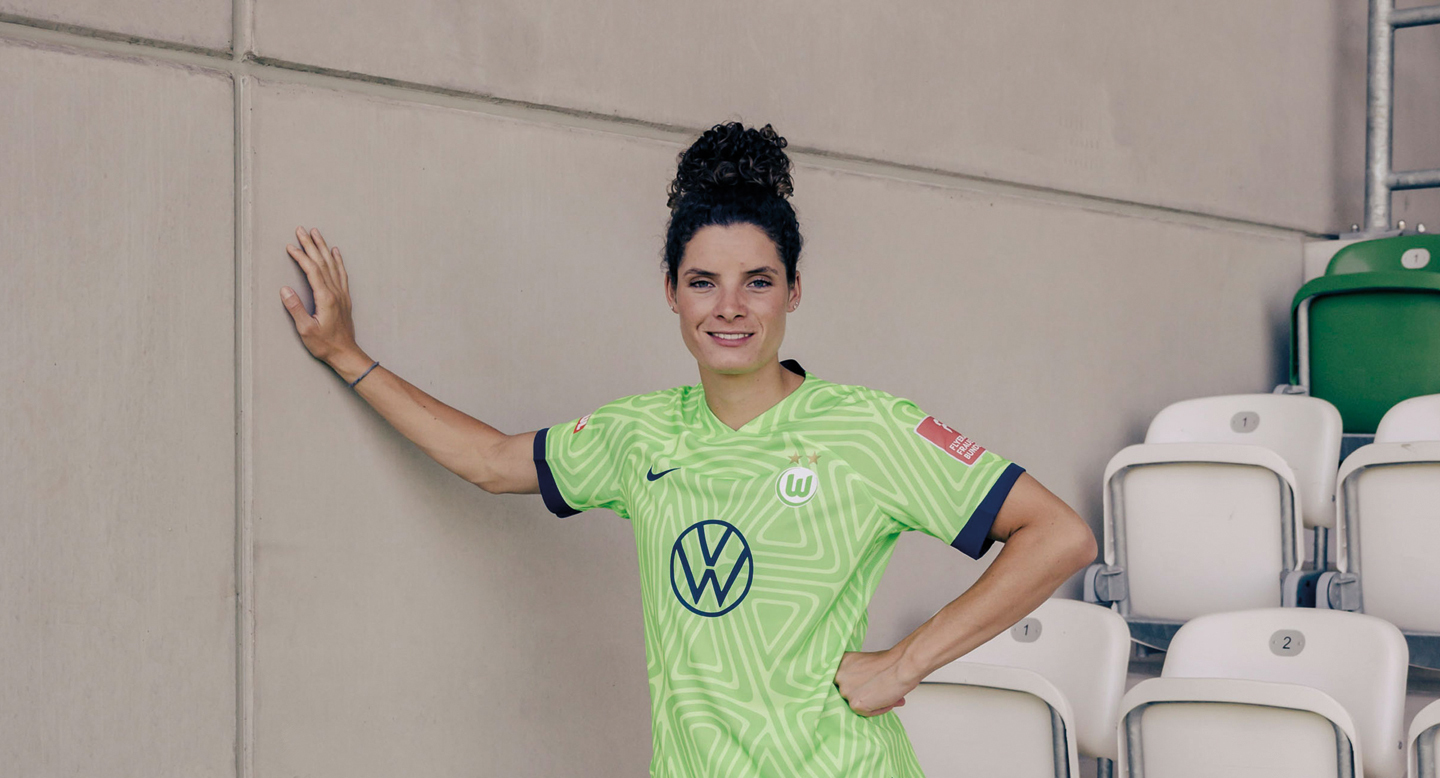 Foto von Dominique Janssen. Die Fußballerin blickt aus einem hellgrünen Trikot lächelnd in die Kamera. Mit einer Hand stützt sie sich an einer Wand.