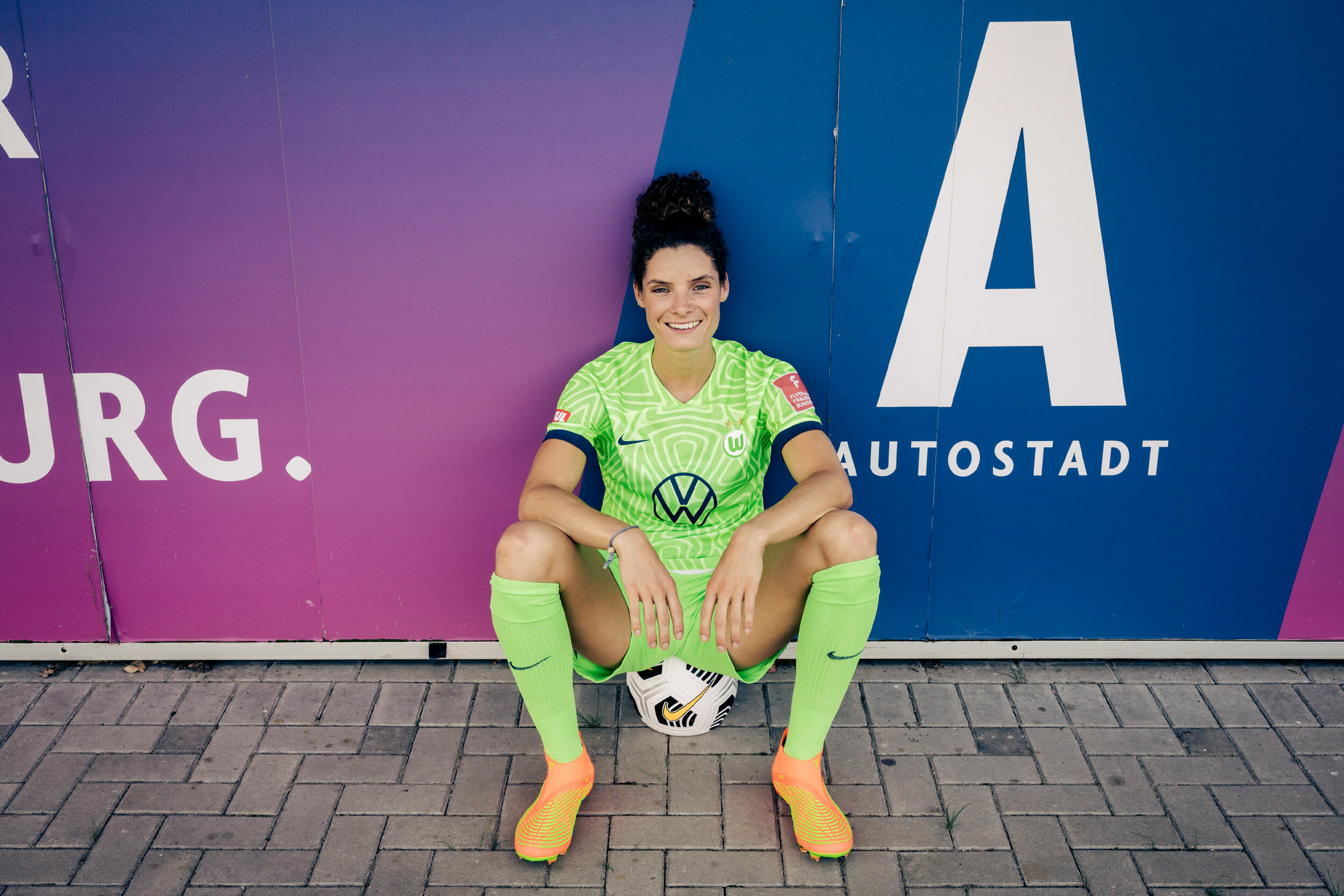 Foto von Dominique Janssen. Die Fußballerin sitzt auf einem Fußball an eine Wand gelehnt, lächelt in die Kamera. Auf der Wand steht Autostadt.