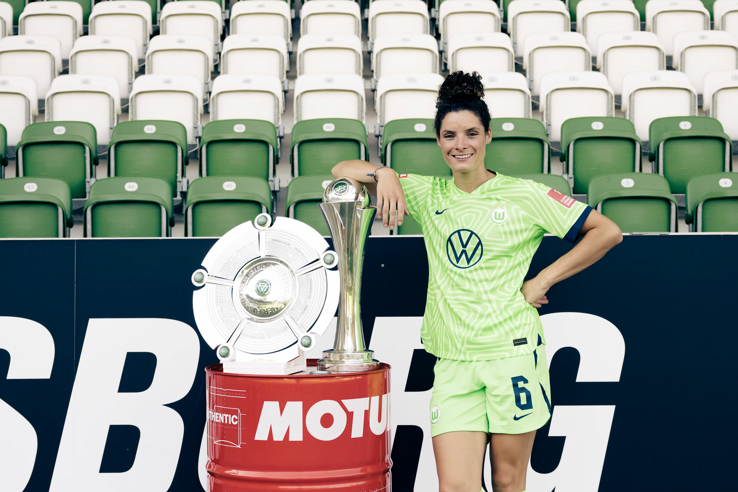 Foto von Dominique Janssen, die an einem Pokal lehnt. Der Pokal steht auf einer roten Tonne. Im Hintergrund sind Stadionsitze in grün und weiß.