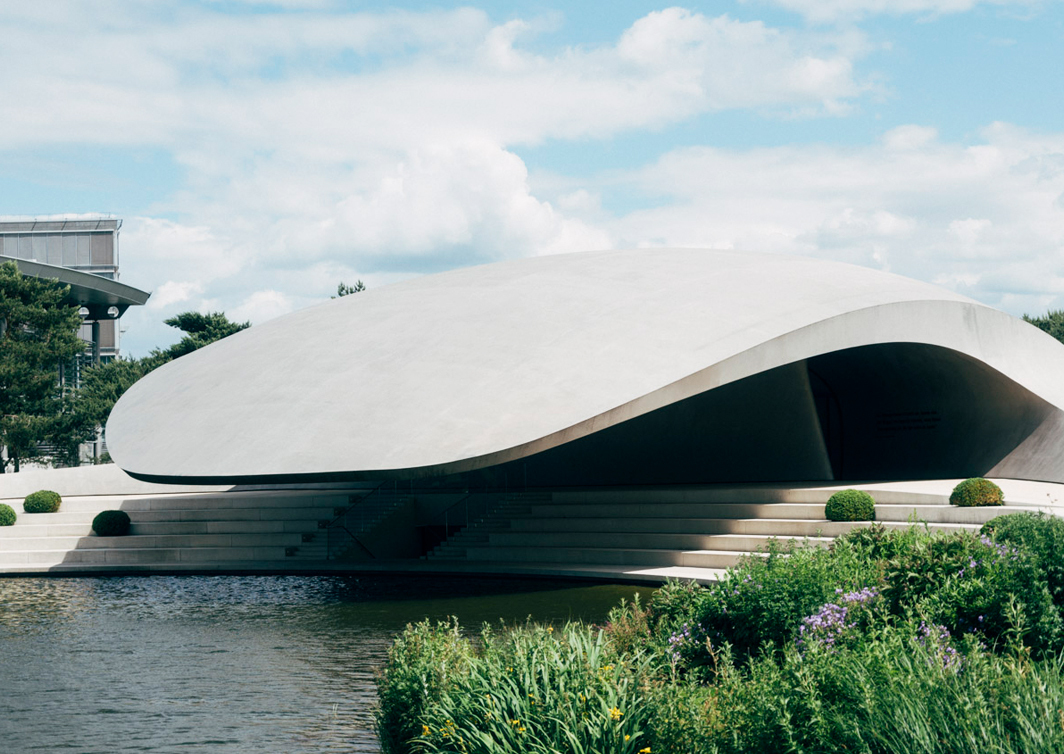 Foto der organischen Architektur des Porsche Pavillon. Ein rundes, geschwungenes Dach bedeckt Treppen an einem Gewässer. Der Himmel ist wolkig-blau.