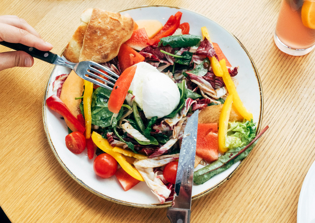 Foto von einem Teller, auf dem bunter Salat, Mozarella und ein Stück Ciabatta liegt.Links vom Teller hält eine Hand eine Gabel, rechts ist ein Messer.