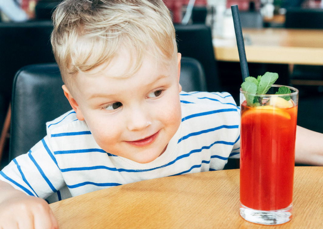 Foto von einem weißen Kind, das an einem Tisch sitzt und eine rote Limo mit Minze und Strohhalm betrachtet und anlächelt.