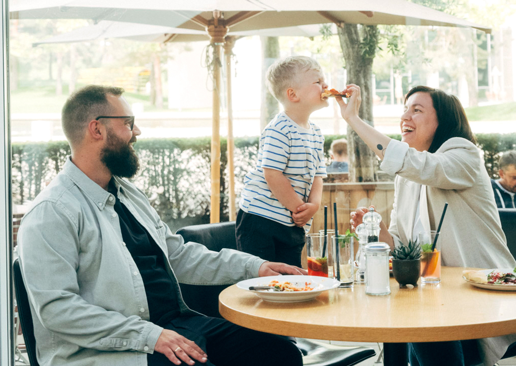 Foto einer Familie, die an einem Tisch sitzt und lacht. Die Frau hält ein Stück Pizza. Das Kind steht auf dem Stuhl und beisst in das Stück Pizza.