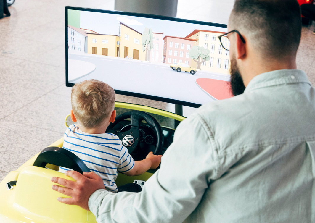 Foto von Vater und Kind in Rückenansicht. Das Kind sitzt in einem Mini-VW vor einem Bildschirm, die Hände am Lenkrad. Der Vater kniet daneben.