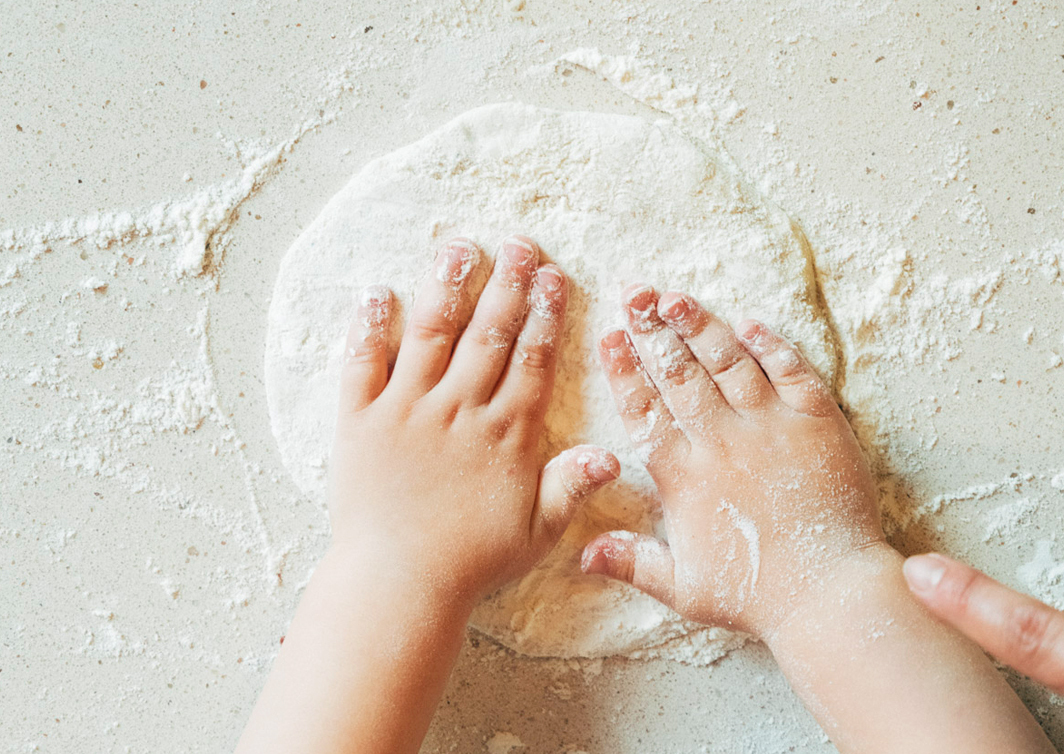 Foto von zwei weißen Kinder-Händen, die auf einem runden Teig liegen. Um den Teig und auf den Händen ist Mehl verteilt.