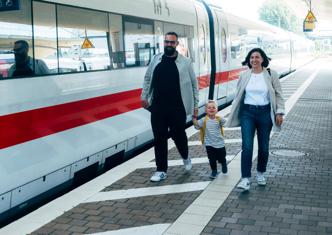 Drei Person laufen neben einem ICE das Bahngleis entlang auf die Kamera zu. Zwei Erwachsene halten in ihrer Mitte ein Kind an den Händen. Alle lachen.