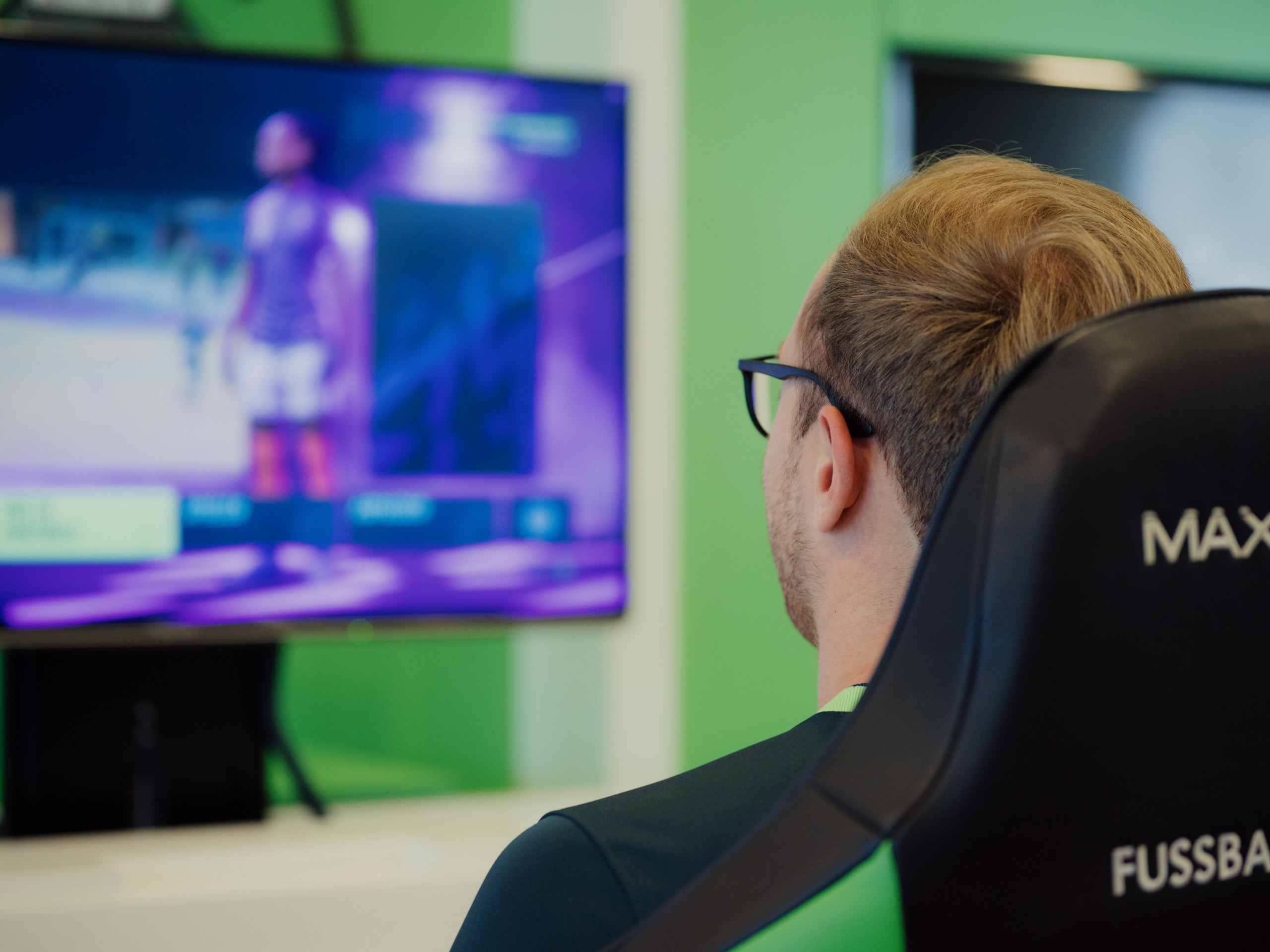 Foto von Max Malle in Rückansicht. Der Gamer sitzt im Stuhl und blickt auf den Bildschirm. Dort steht ein Mensch im Trikot.