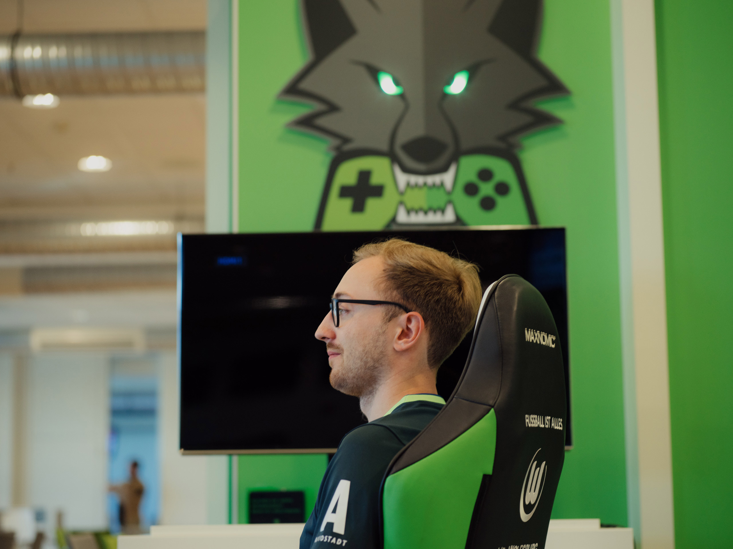 Foto von Max Malle im Profil. Der Gamer sitzt im Stuhl, im Hintergrund befindet sich ein Bildschirm und ein illustrierter Wolf mit Controller im Mund.