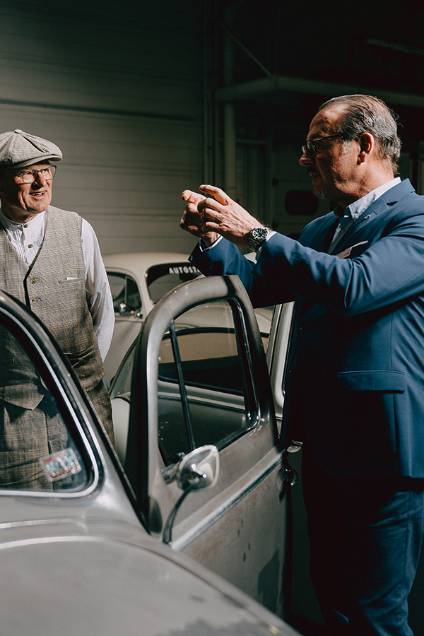 Foto zweier Personen,die in einer Ausstellungshalle über eine geöffneten Autotür miteinander kommunizieren. Eine Person gestikuliert,die andere lacht.