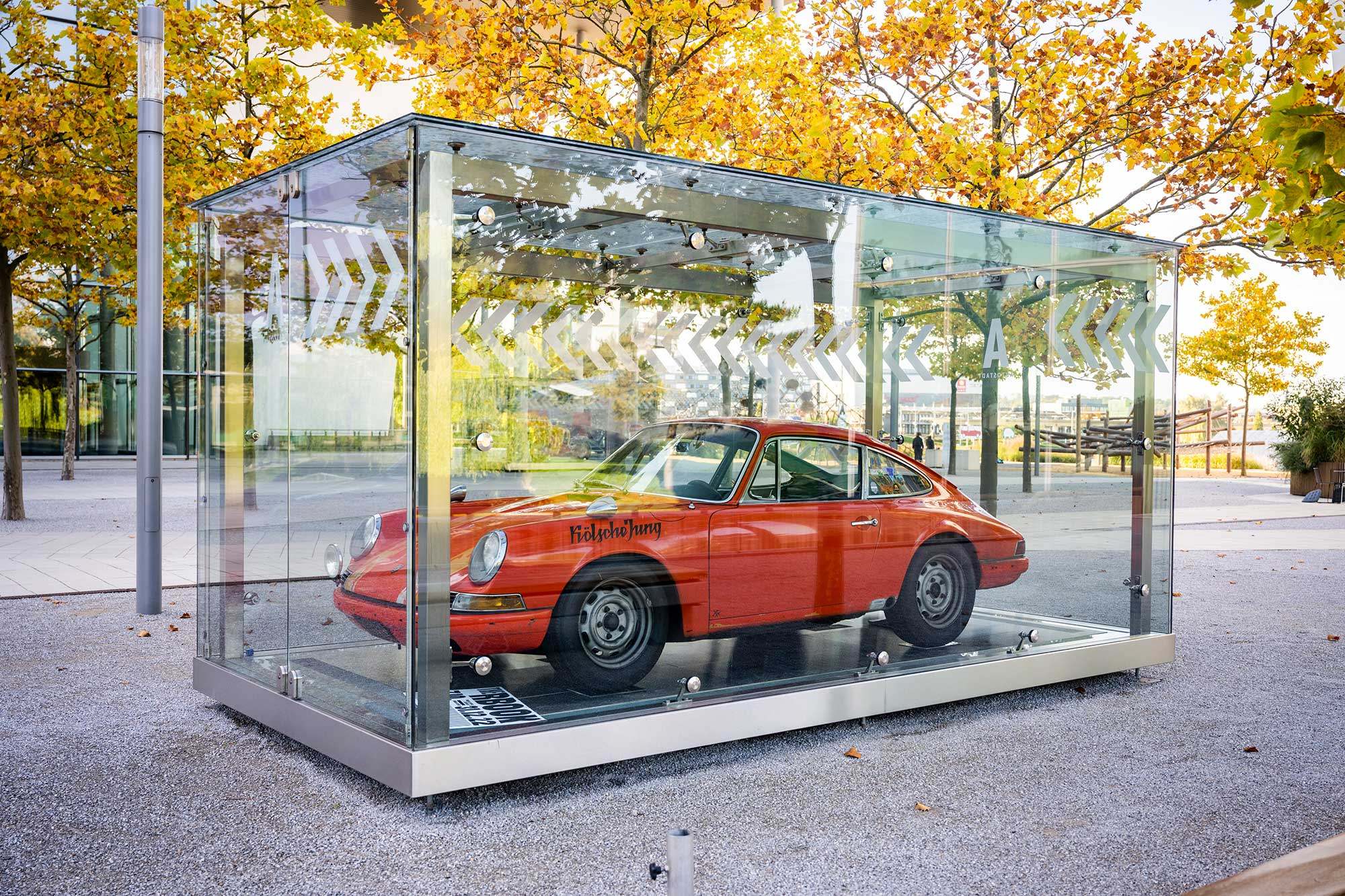 Foto eines roten Porsche in einem Glaskasten auf dem Autostadtgelände umringt von Bäumen. Auf dem linken Kotflügel steht "Kölscher Jung".