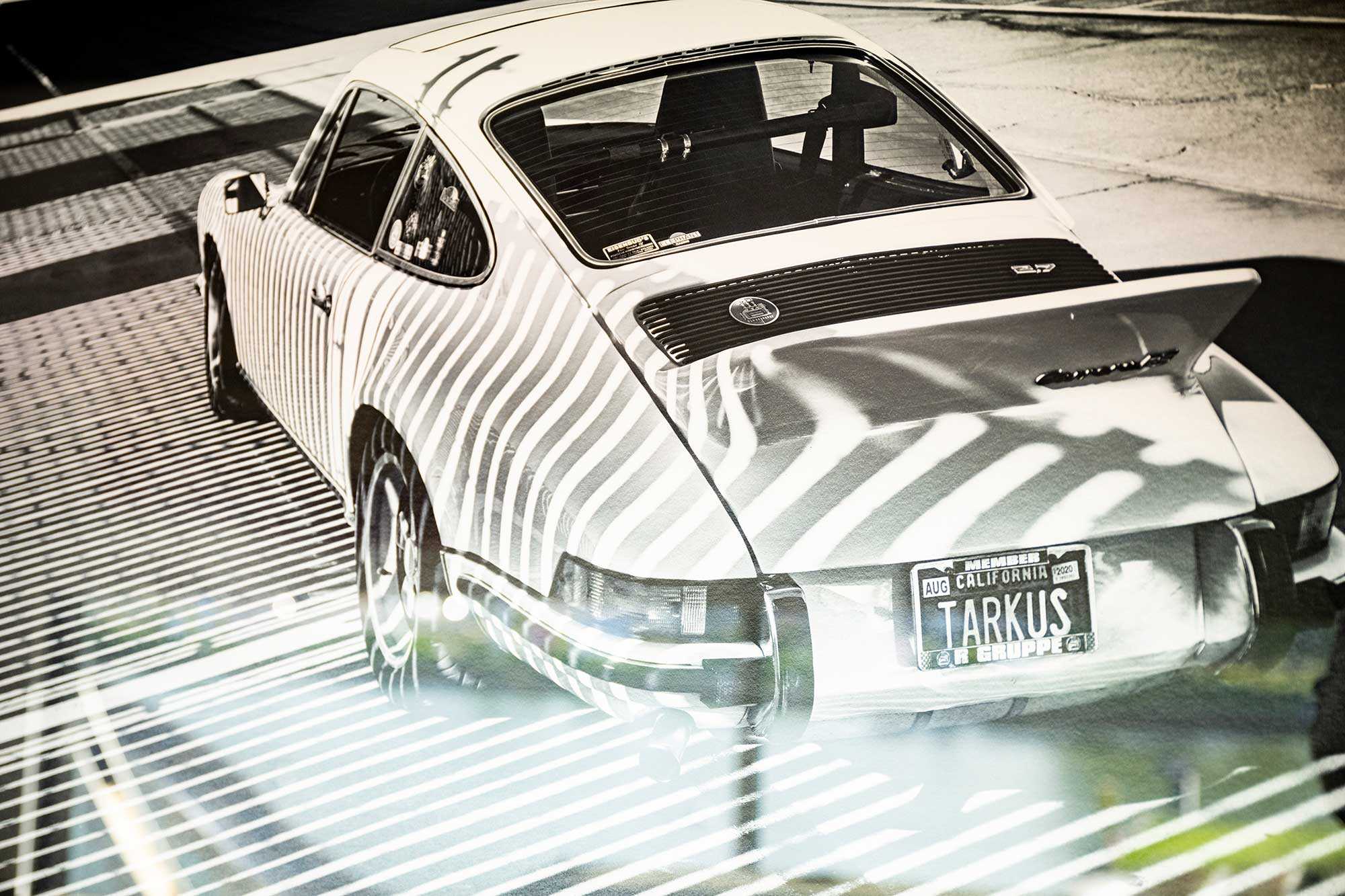 Nahaufnahme einer Schwarzweißfotografie eines weißen Porsche im Schatten eines Gitters. Auf dem Nummerschild steht "TARKUS".