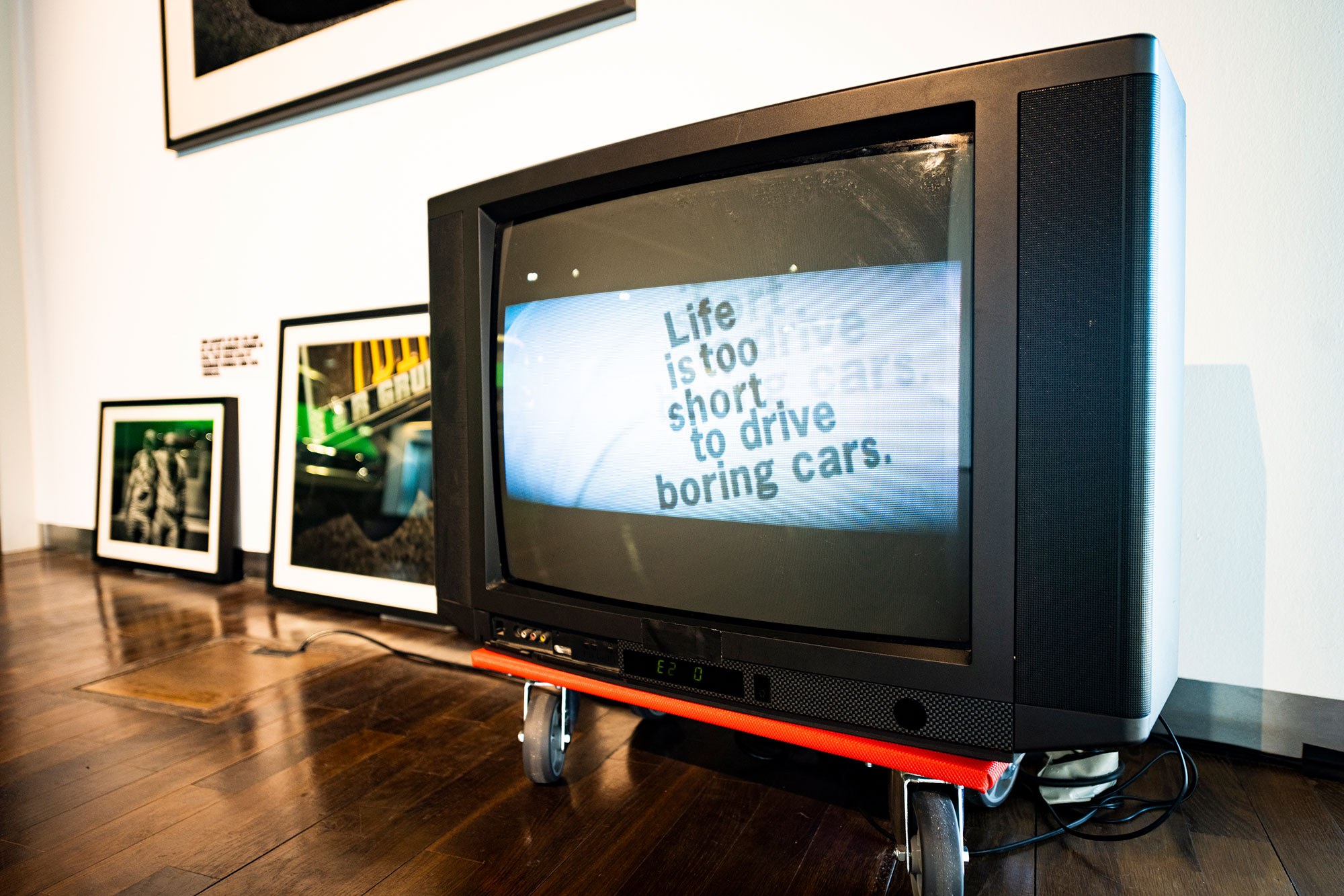 Fotografie eines Fernsehapparats auf einem Rollbrett in einem Ausstellungsraum. Links daneben lehnen zwei gerahmte Fotografien an einer weißen Wand.