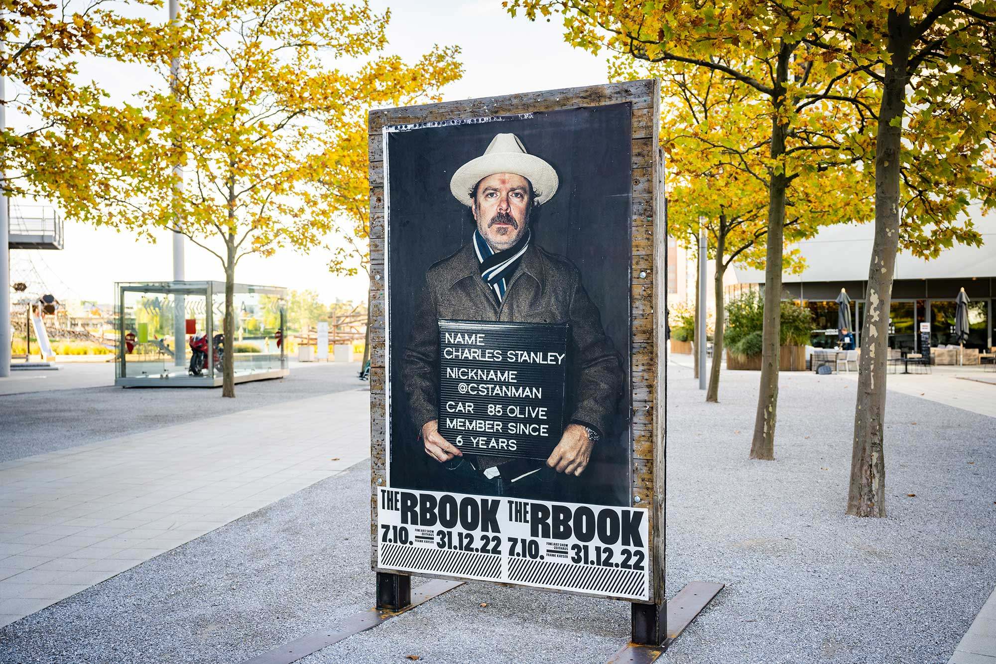Foto eines Poster-Aufstellers auf dem Autostadt-Gelände umringt von Bäumen. Auf dem Poster ist eine Person mit Bart, Cowboyhut, ein Schild haltend.