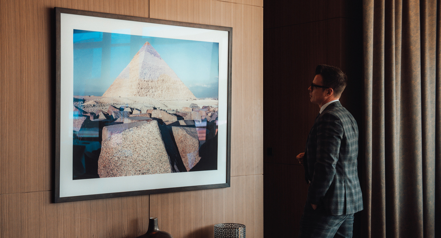 Foto eines Raumes, in dem ein großes gerahmtes Foto einer Pyramide an einer Holzwand hängt. Rechts steht ein Mensch im Anzug, der das Bild anblickt.