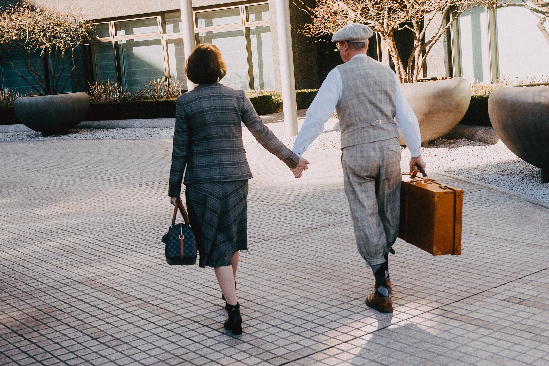 Foto von zwei Personen in der Rückansicht. Die beiden laufen Hand in Hand,mit Koffern in der anderen Hand, in historischen Outfits auf ein Gebäude zu.