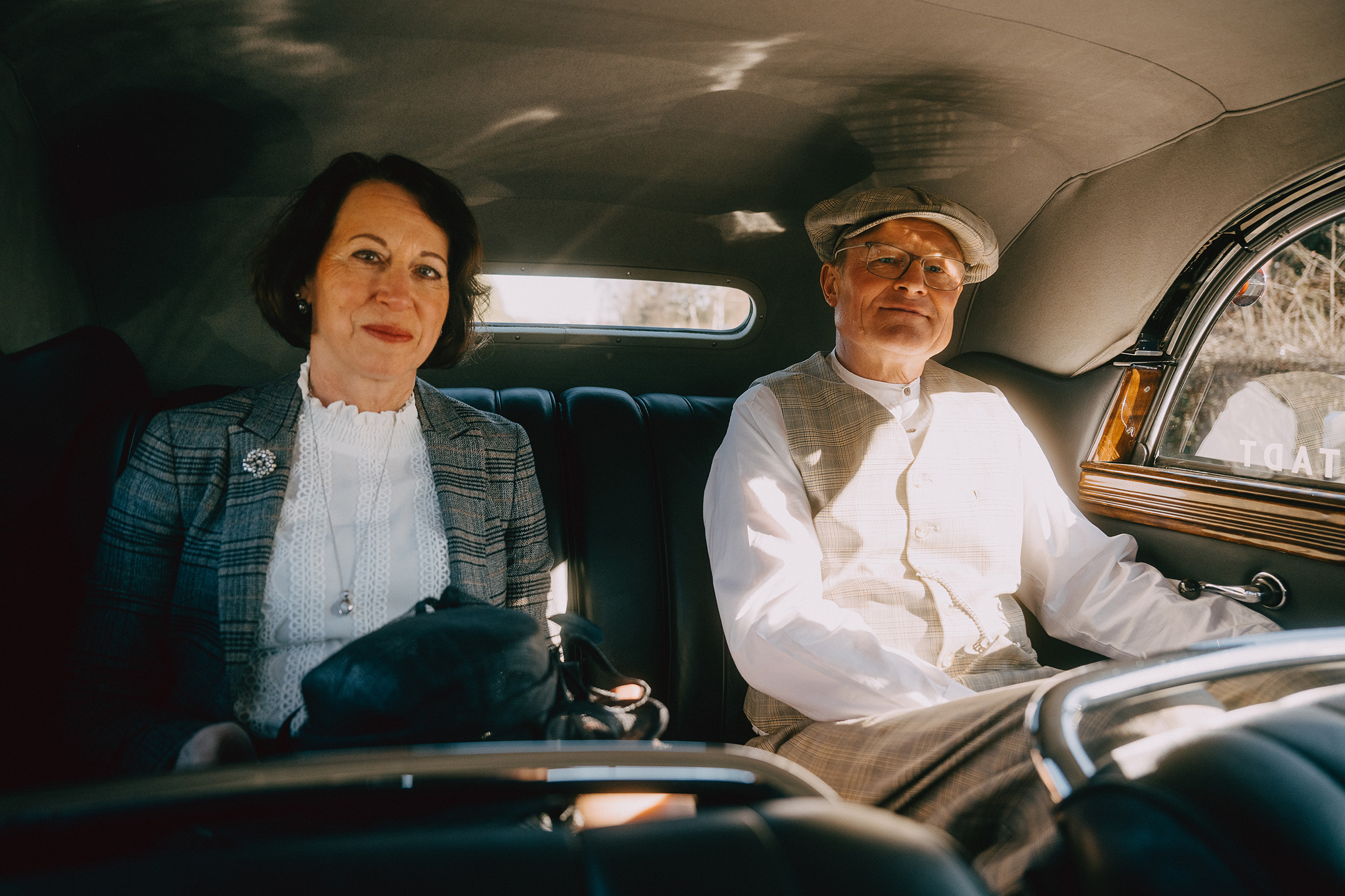 Foto von zwei Personen, die in einem historischen Cabrio sitzen. Das Dach ist geschlossen. Die beiden blicken aus historischen Outfits in die Kamera.
