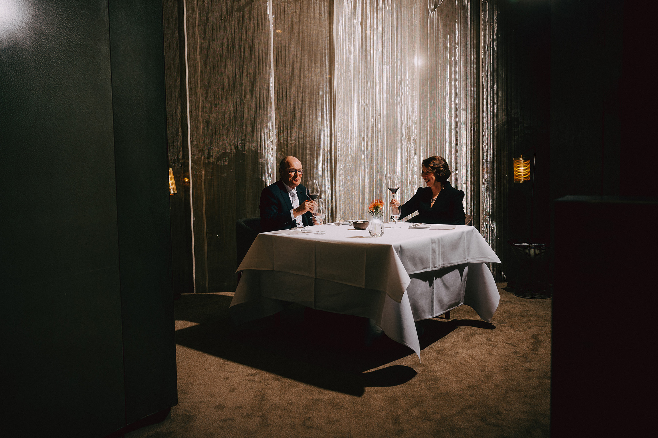 Foto von zwei Personen an einem gedeckten Tisch vor einem silbernen Vorhang. Die beiden heben ein Rotweinglas und blicken einander an.