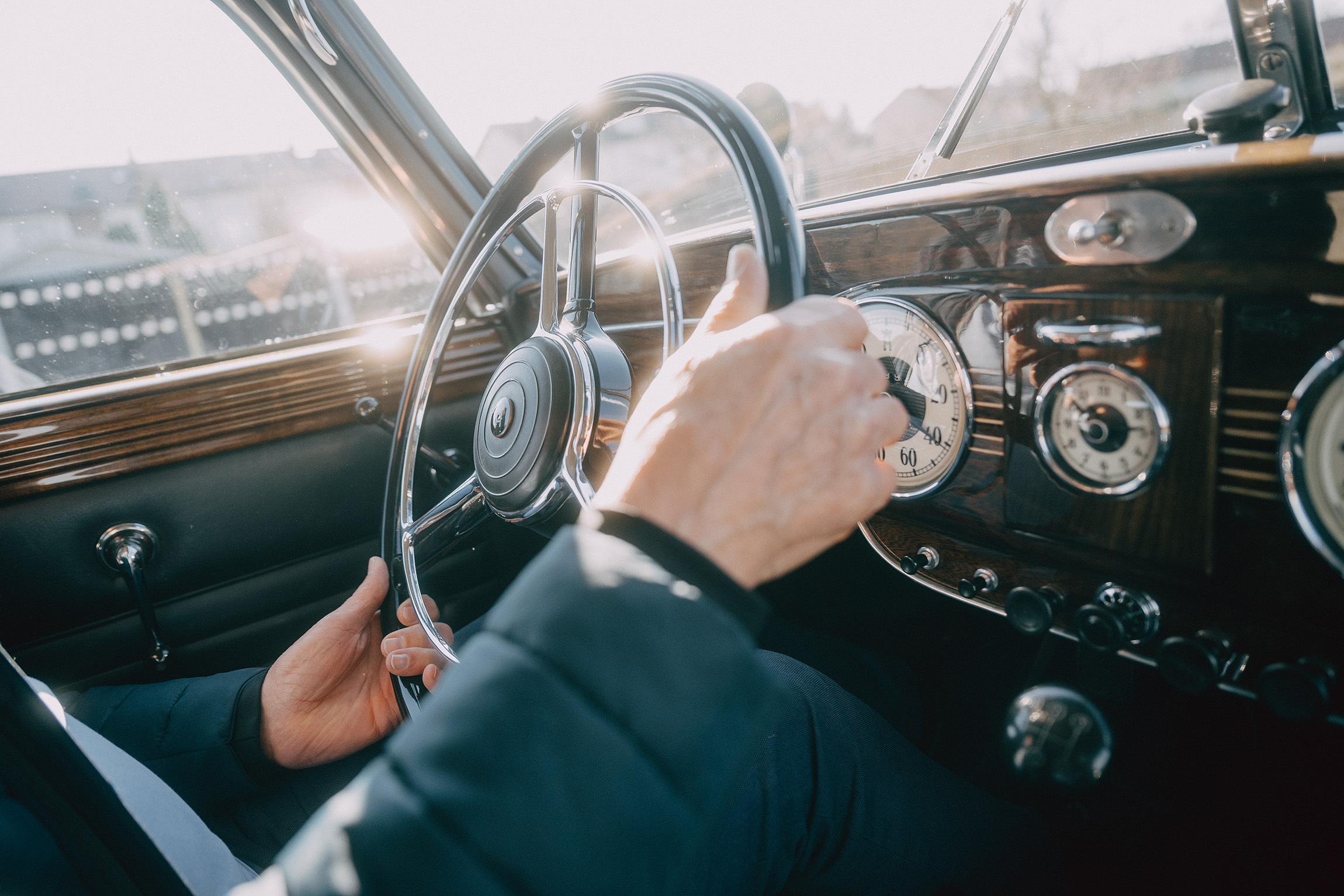 Foto von Händen, die das Lenkrad eines historischen Autos halten. Sonne scheint durch die Fensterscheiben auf die Hände.