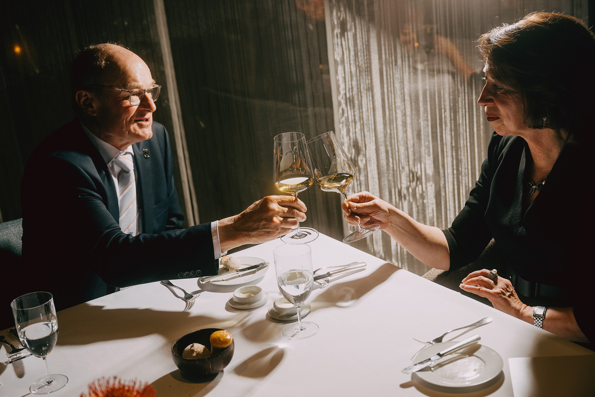 Foto zweier Personen, die an einem gedeckten Dinner-Tisch sitzen und mit den Weingläsern in ihrer Hand anstoßen.