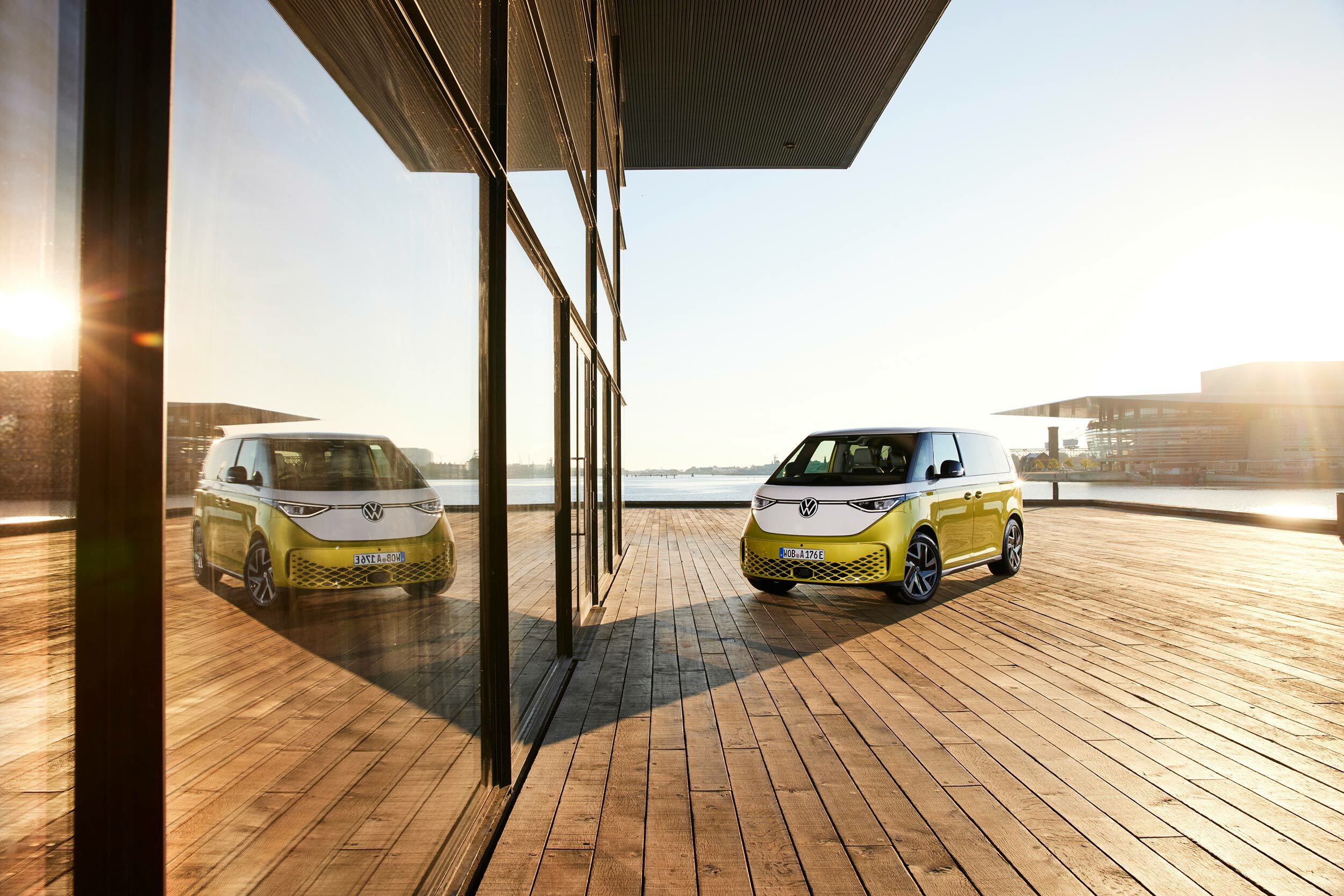 Foto eines parkenden weiß-goldenen VW ID Buzz. Das Auto parkt neben einem Gebäude mit gläserner Fasade, in dem sich das Auto spiegelt.