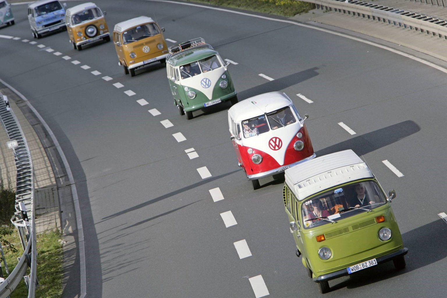 Fotografie von sechs bunten VW Bullis, die in einer Reihe auf einer Autostraße hintereinander fahren.